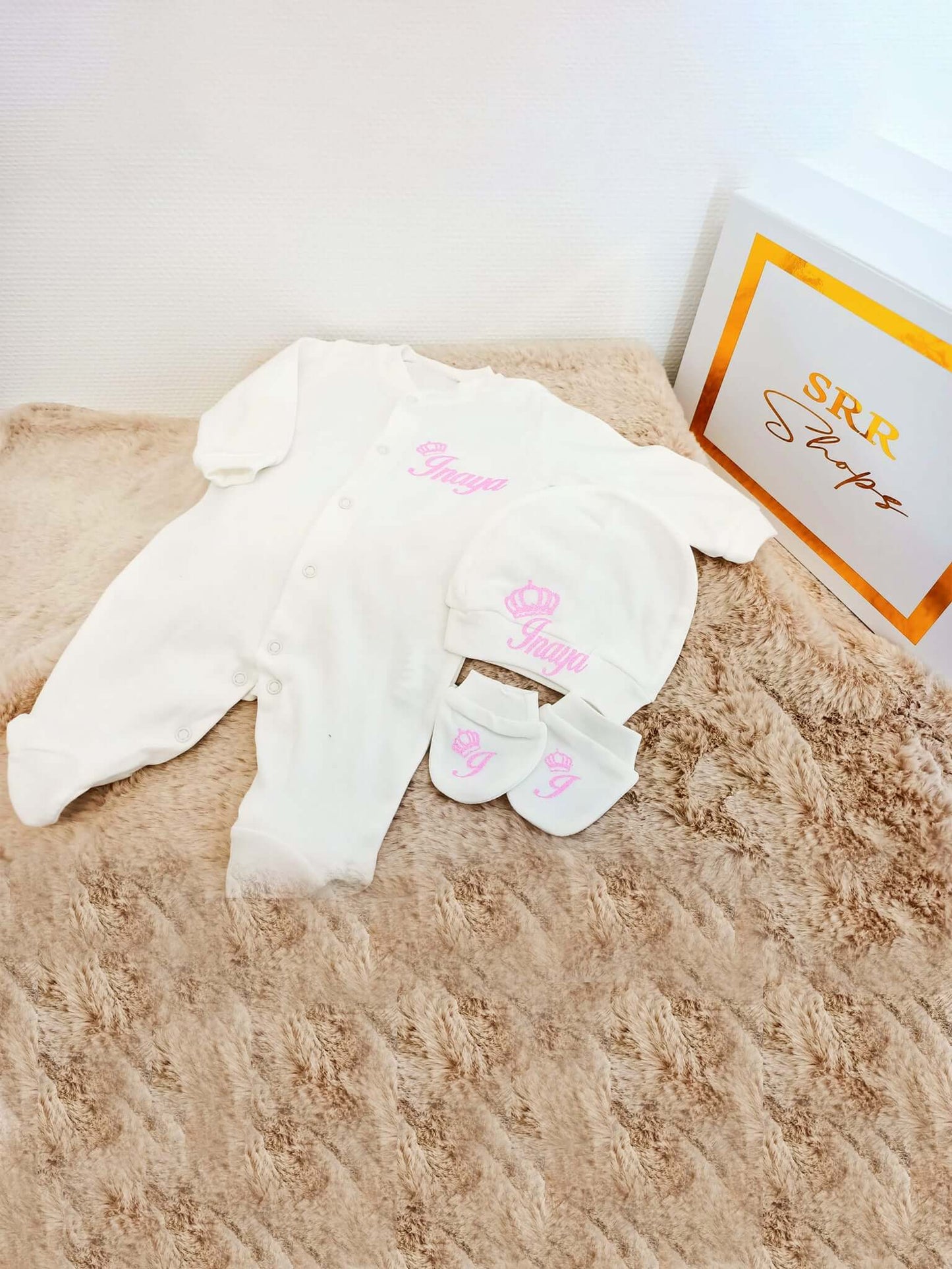 Pyjamas bébé personnalisés blancs avec bonnet et chaussons roses sur un lit beige.